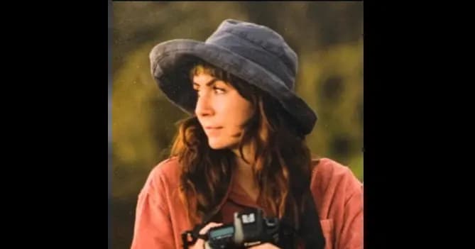 Jessi is standing in a field, wearing a hat and a corduroy shirt, holding a camera and looking to the right. 
