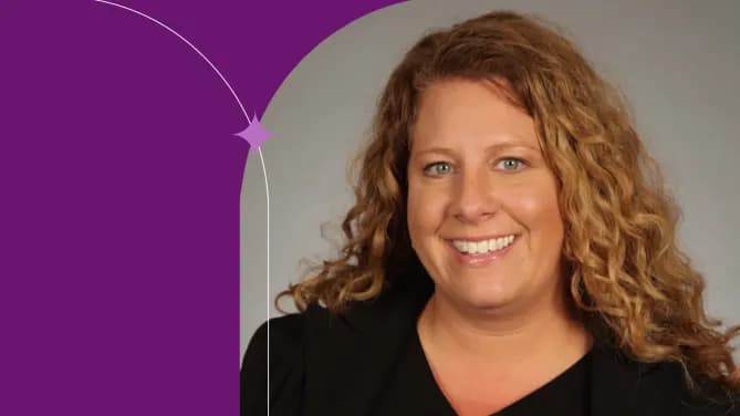 Leila has curly strawberry blonde hair and is wearing a black v-neck shirt. She is sitting in front of a grey background smiling for the camera.