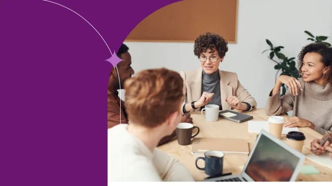 Five people sit at a table talking. The person in the middle has short hair and wears a turtleneck and blazer. The person on the right wears a sweater, and the people on the left face away from the camera. One has red hair, the other has short black hair.